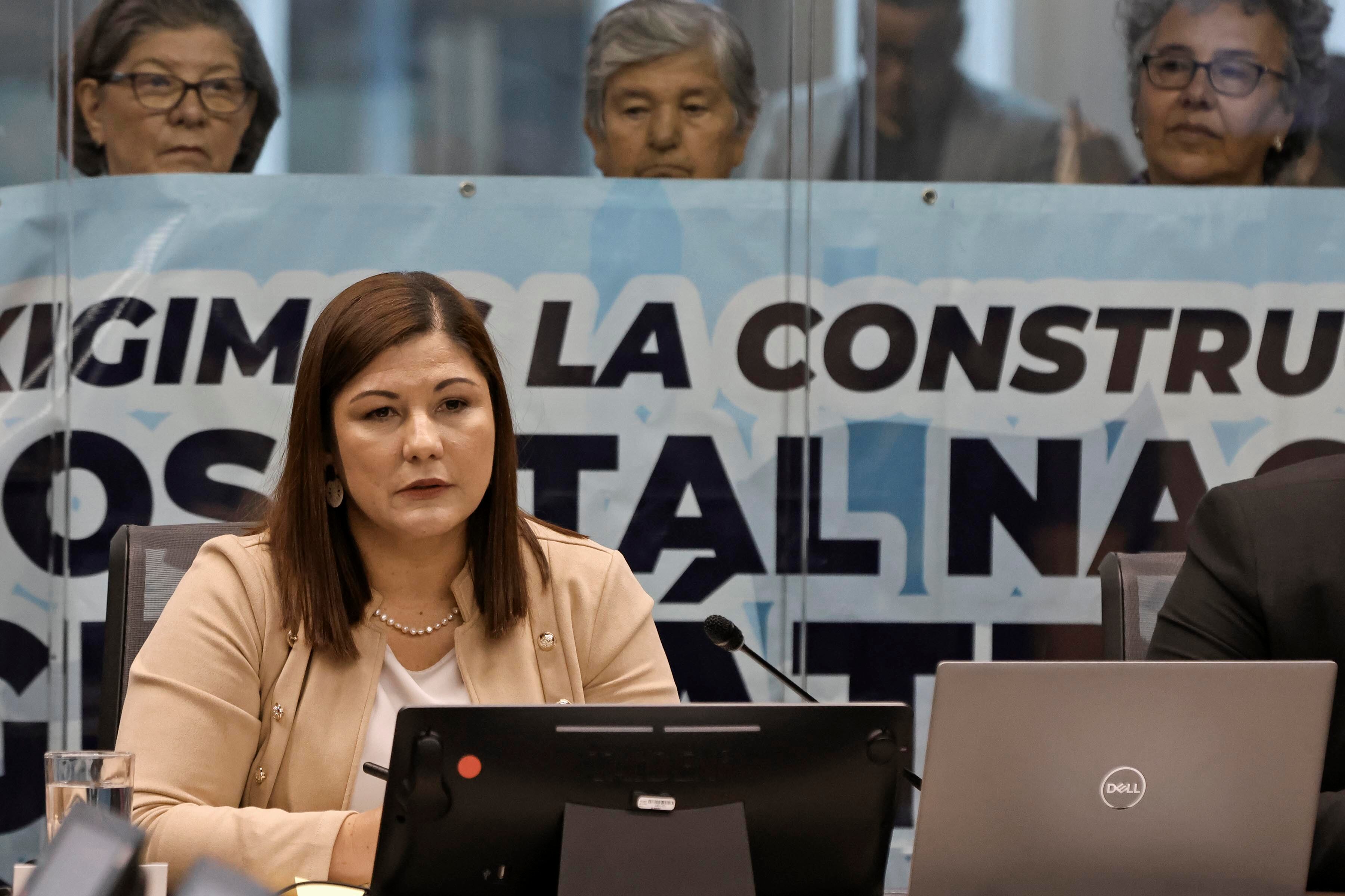 Mujer joven, pelo oscuro a los hombros, vestida con chaqueta color papaya. Detrás de ella, tras una vidriera, personas con una manta.