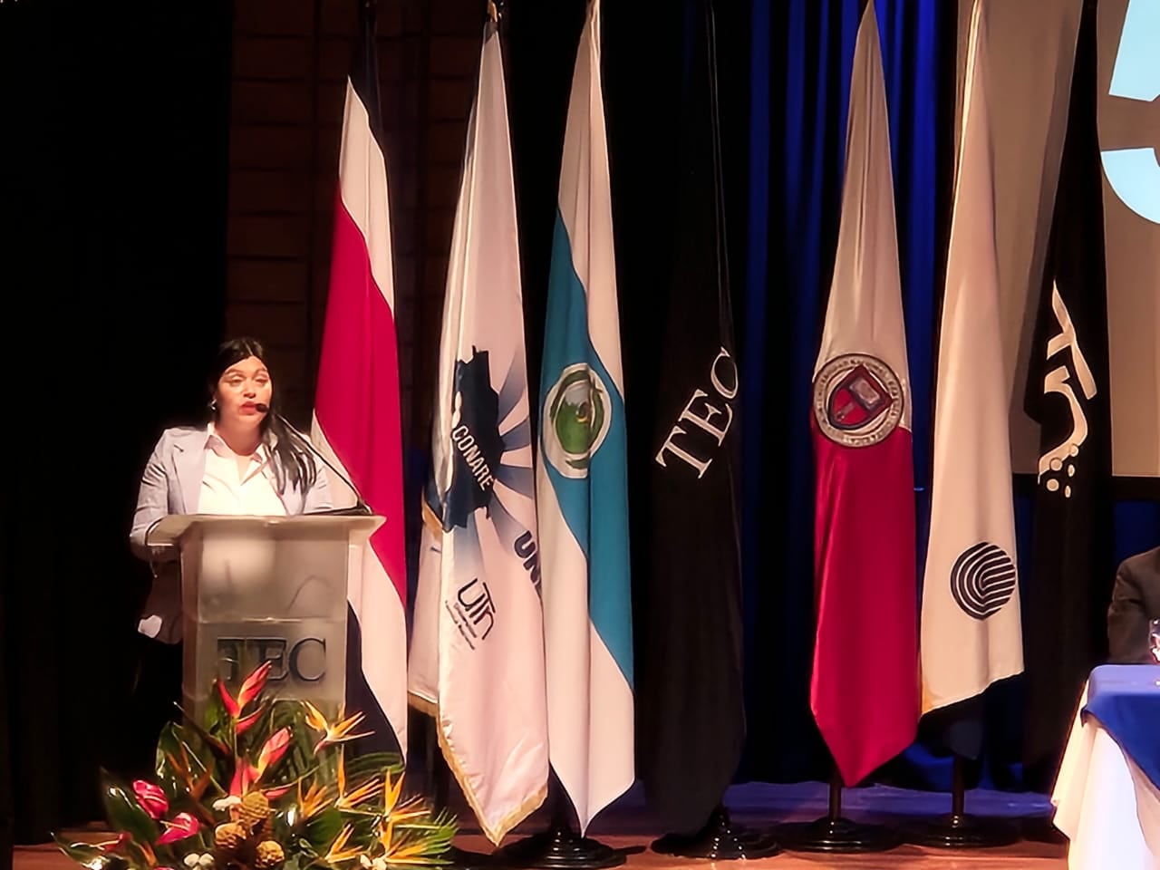 María Estrada, rectora del Tec da su discurso en un podio en un escenario donde se ven las banderas de las universidades públicas.