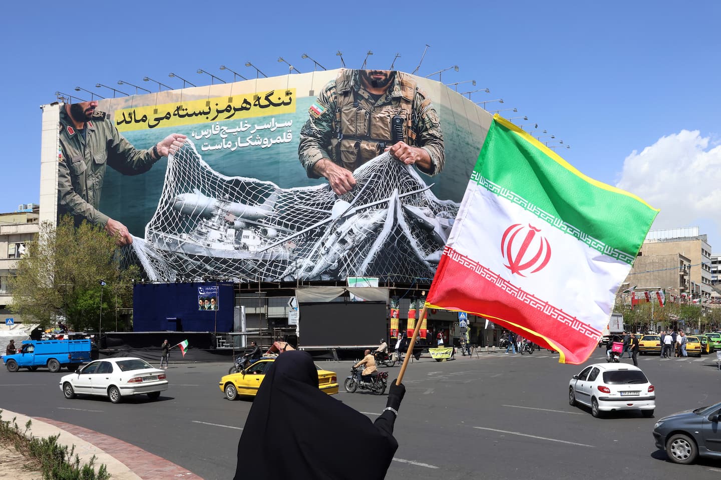 Una mujer sostiene la bandera nacional de Irán mientras está de pie cerca de una valla publicitaria con la frase "El estrecho de Ormuz permanece cerrado" en la plaza Enqelab de Teherán, el 5 de abril de 2026. Omán e Irán mantuvieron conversaciones para facilitar el paso por el estrecho de Ormuz, según informó la agencia estatal de noticias omaní el 5 de abril, ya que este punto estratégico para el transporte marítimo se encuentra prácticamente cerrado debido a la guerra en Oriente Medio.