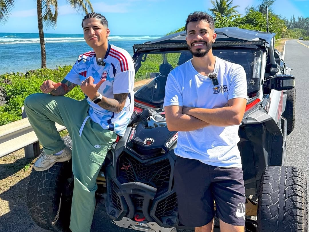 Julián Valverde a la izquierda y Araya Vlogs a la derecha, ambos apoyados en un carro durante un viaje a Puerto Rico.