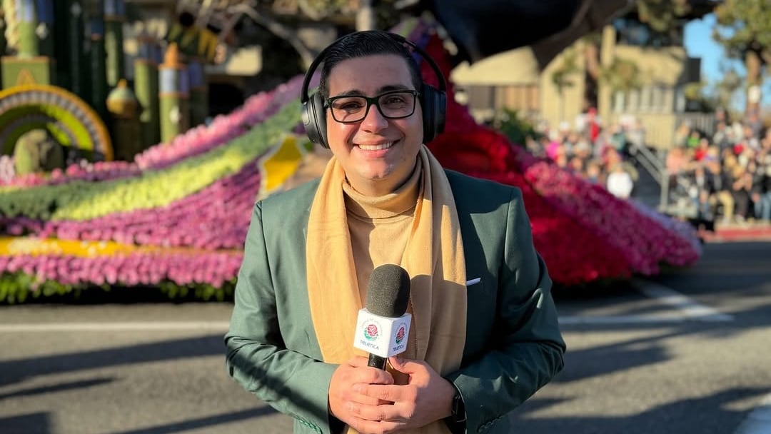 El periodista Elías Alvarado durante una transmisión, sostiene un micrófono en las manos, usa traje verde y una bufanda amarilla.