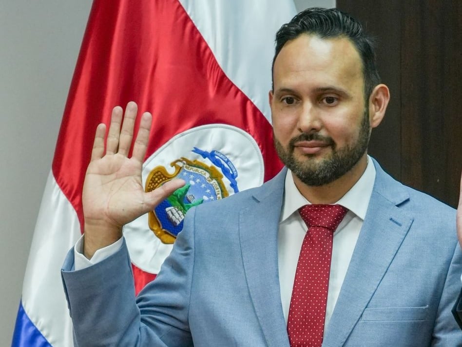 Se observa un hombre con traje entero con la mano derecha levantada para ser juramentado. Al fondo, la bandera de Costa Rica