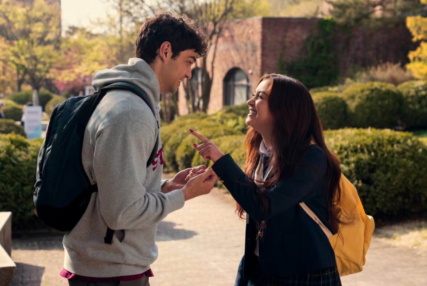 Noah Centineo y Anna Cathcart actuando en la serie 'Besos, Kitty' de Netflix. Ambos aparecen con mochilas escolares.