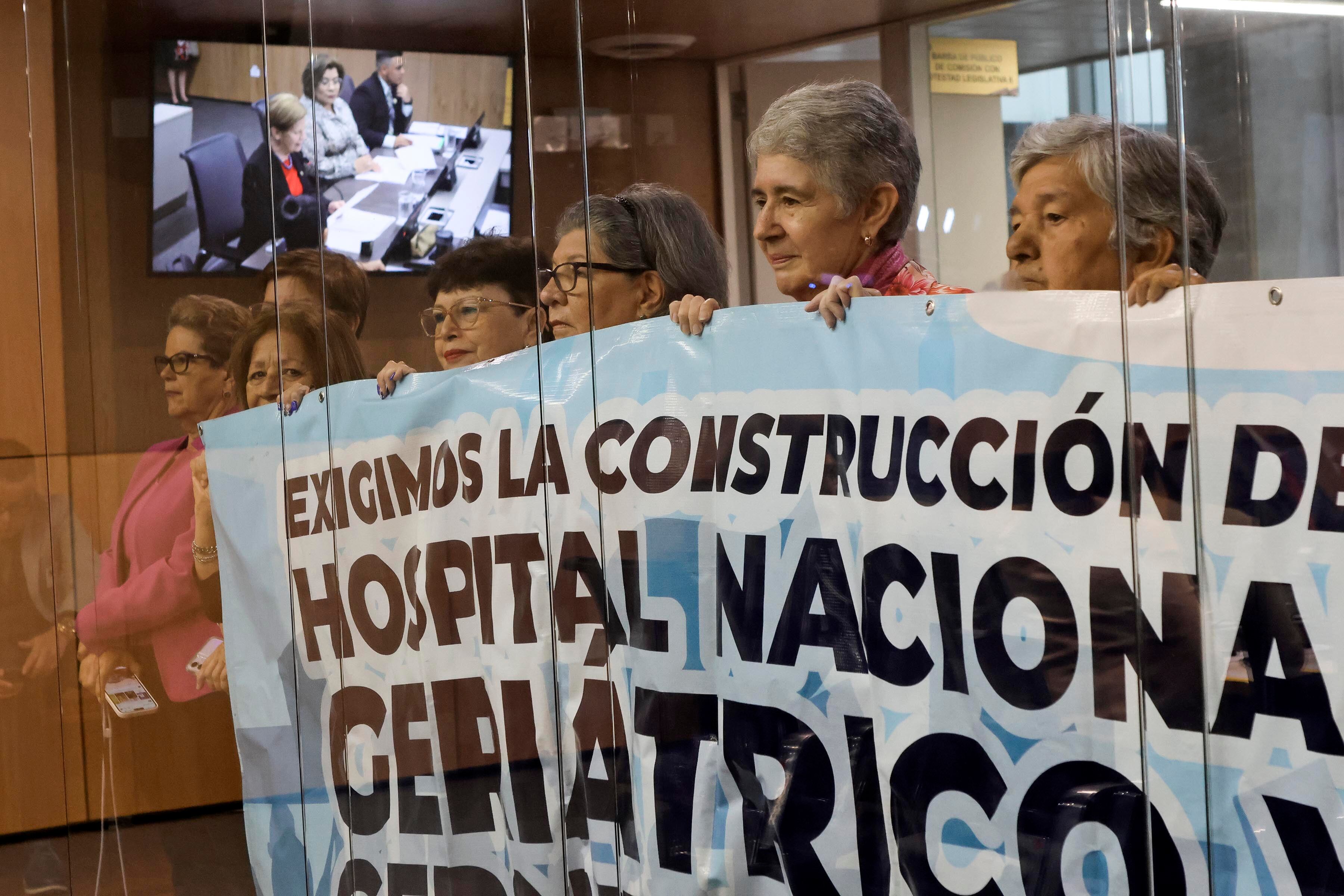 Personas adultas mayores sostienen una manta que pide construir un nuevo hospital.