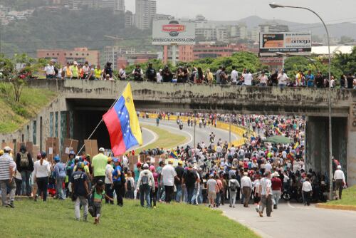 Decenas de venezolanos debajo y sobre un puente en Caracas, uno de ellos con una bandera de su país, ilustrando el valor de los ciudadanos en Venezuela.