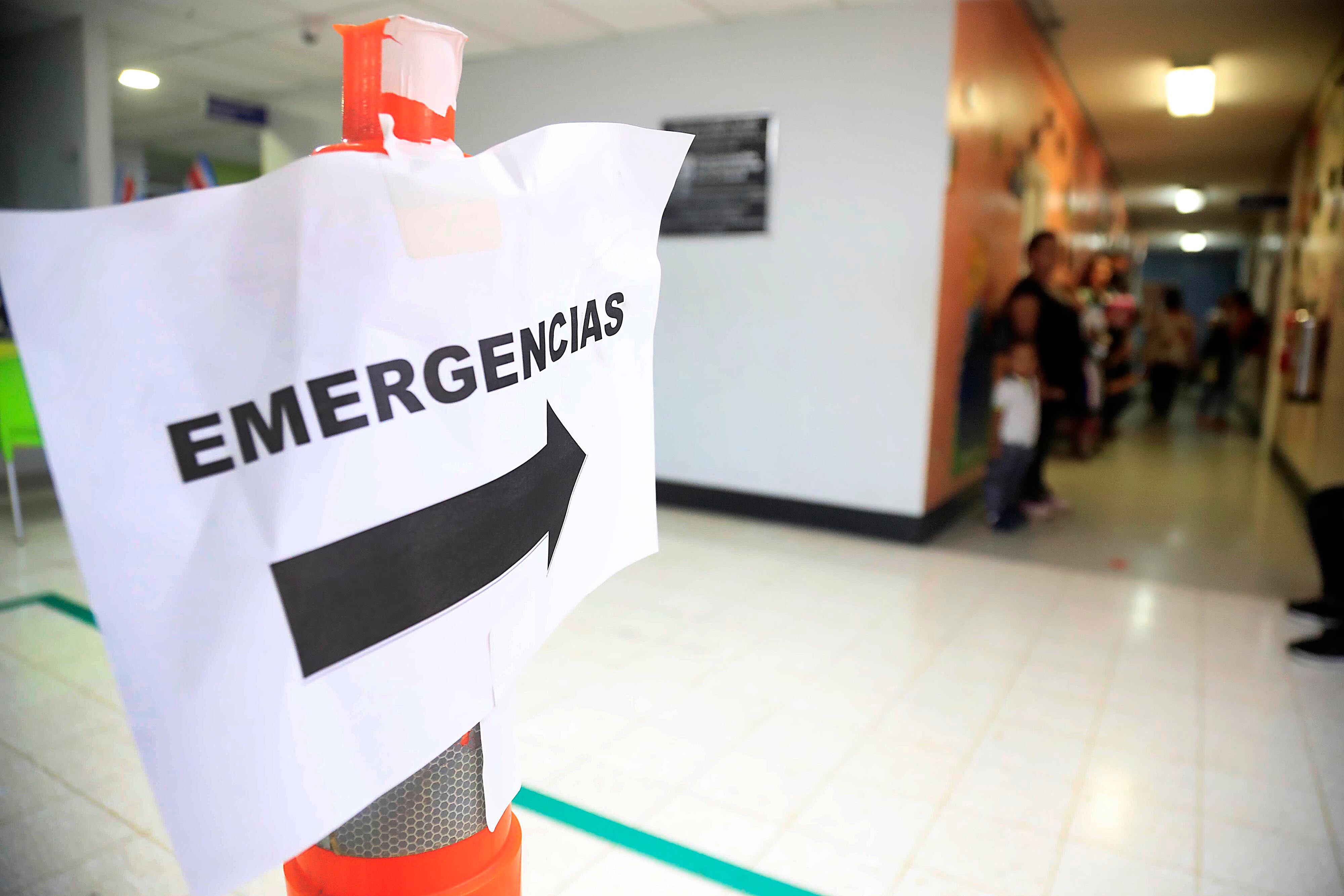 Imagen de un rótulo que dice Emergencias, con una flecha negra que orienta hacia el servicio de emergencias en un hospital. Al fondo, se ve un pasillo lleno de personas, iluminado levemente.