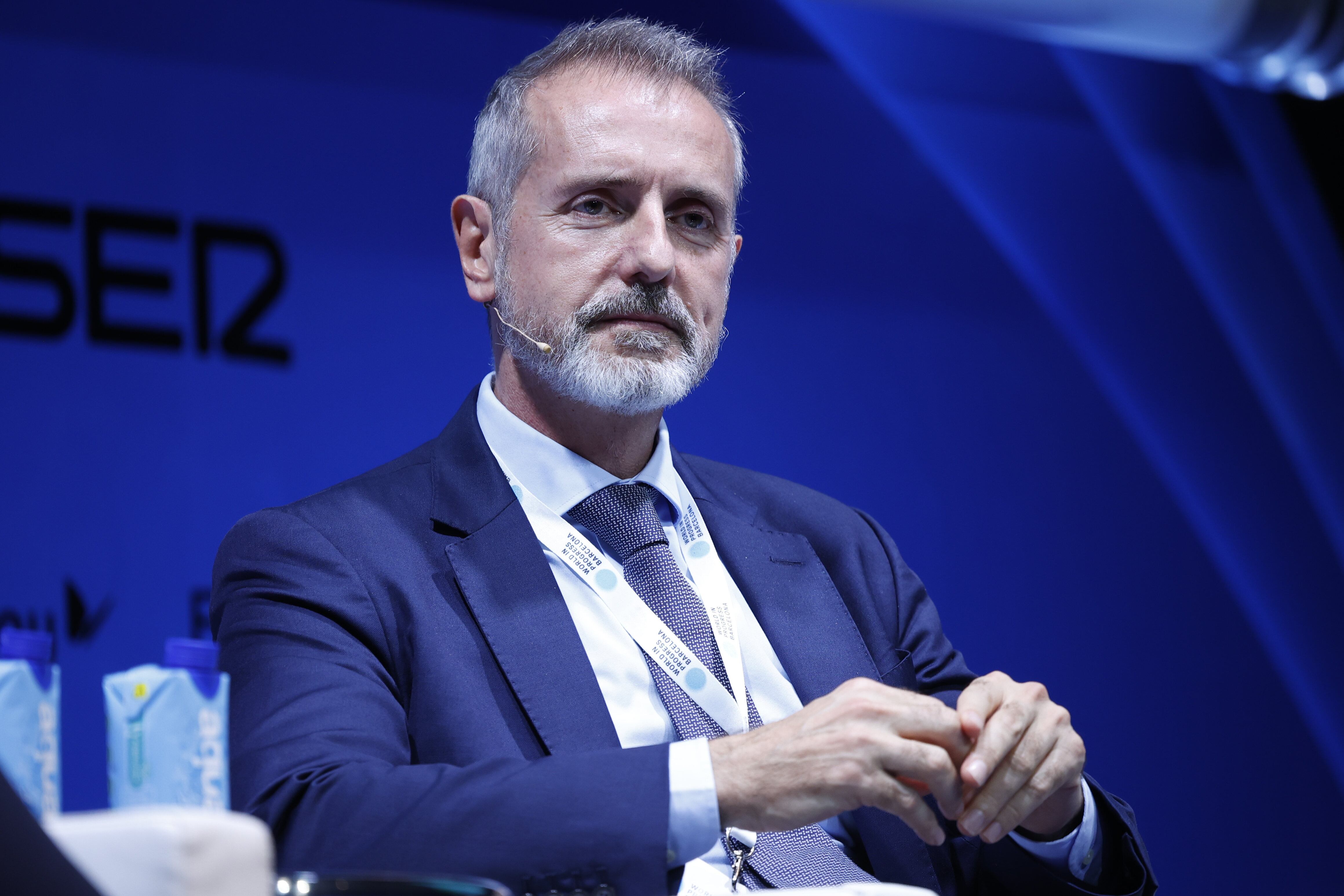 Marc Murtra, con traje azul y corbata, sentado en un escenario con fondo azul durante un evento empresarial, designado recientemente como nuevo presidente de Telefónica.
