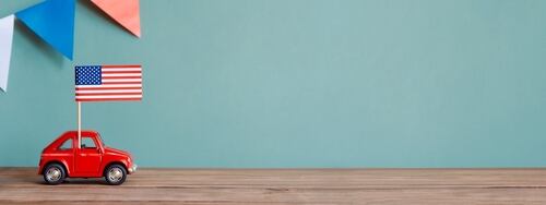 Un auto rojo de juguete con una bandera de Estados Unidos en su techo, sobre una base de madera y un fondo verde con banderines en tonos rojo, blanco y azul.