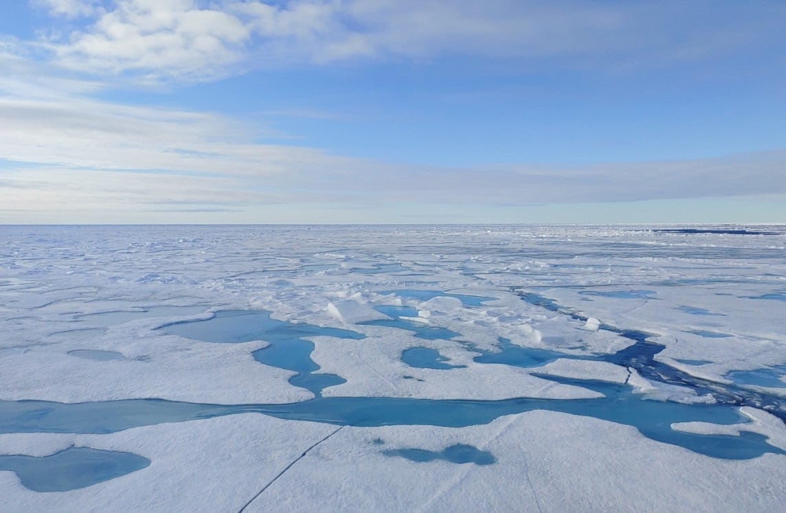 Vista aérea de la capa de hielo marino en el Ártico con grandes grietas y charcos de agua de deshielo, bajo un cielo parcialmente nublado.