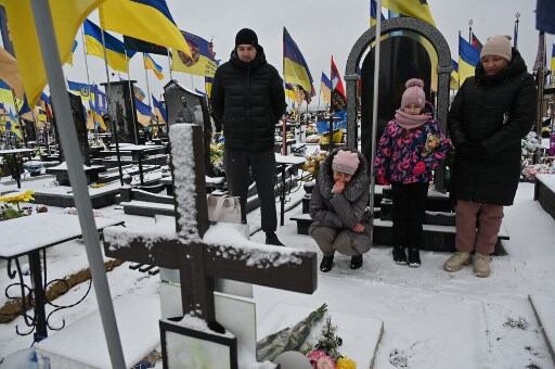 Familia ucraniana en un cementerio cubierto de nieve, rodeados de banderas y tumbas, recordando a sus seres queridos.