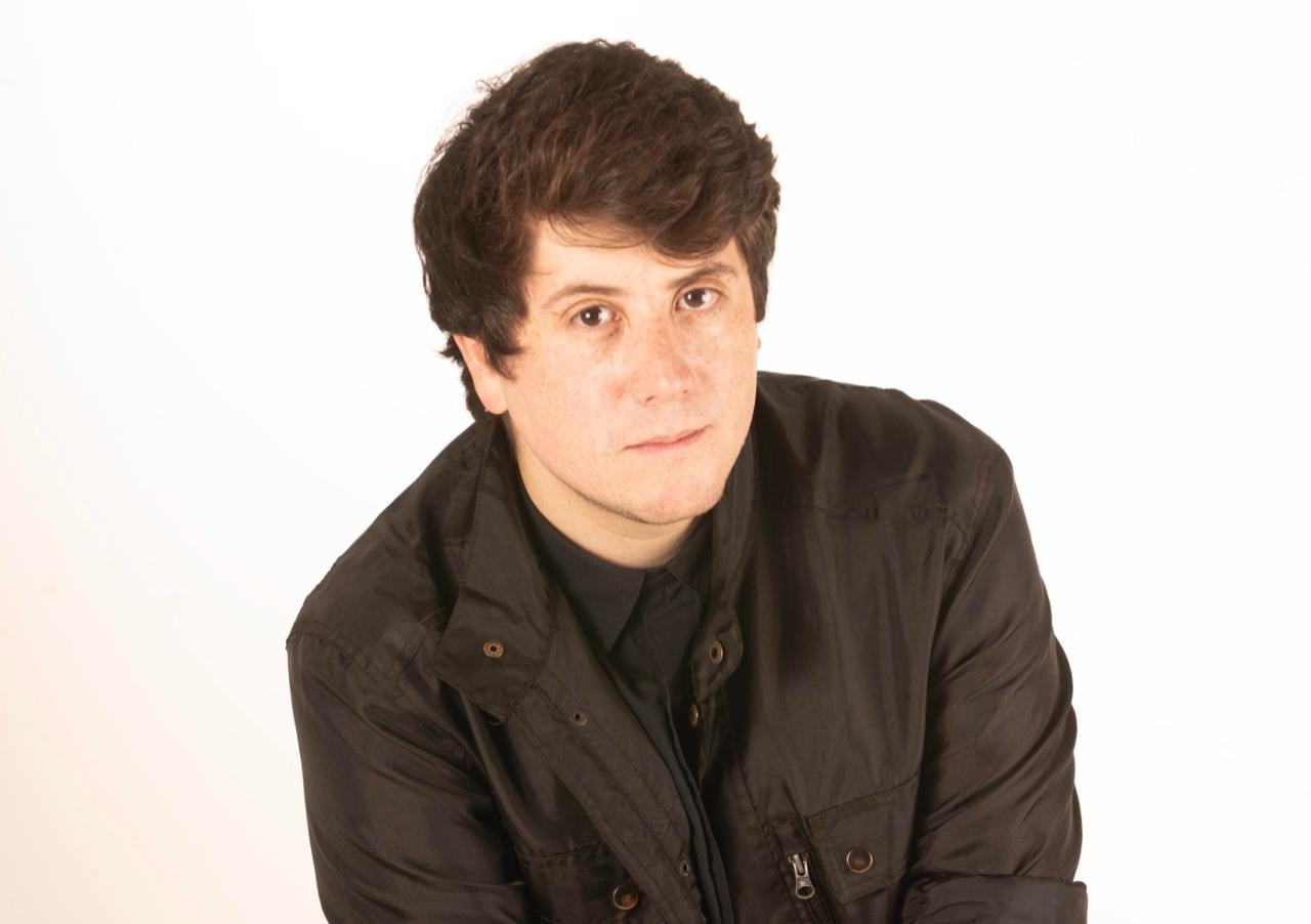 Retrato de hombre joven, vestido de oscuro, en fondo blanco. Leve sonrisa en su rostro.