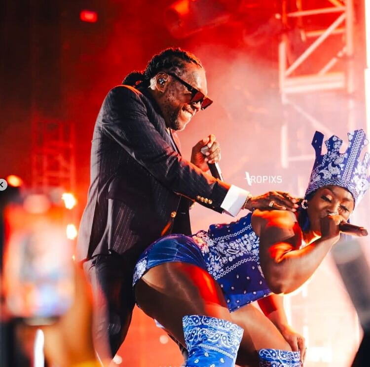 Vybz Kartel cantando en concierto, con el micrófono en mano. usa un traje de color negro y corbata dorada.