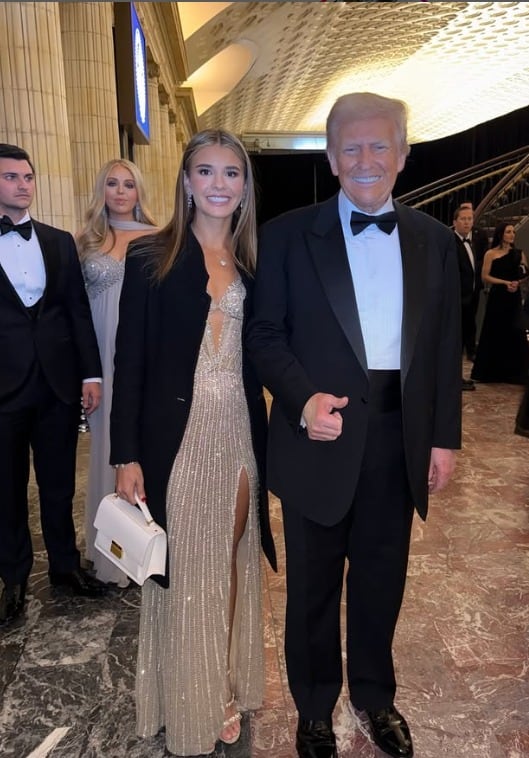 Kai Trump posando con su abuelo Donald Trump. Ella lleva un vestido largo en tono dorado, él viste traje negro con camisa blanca.