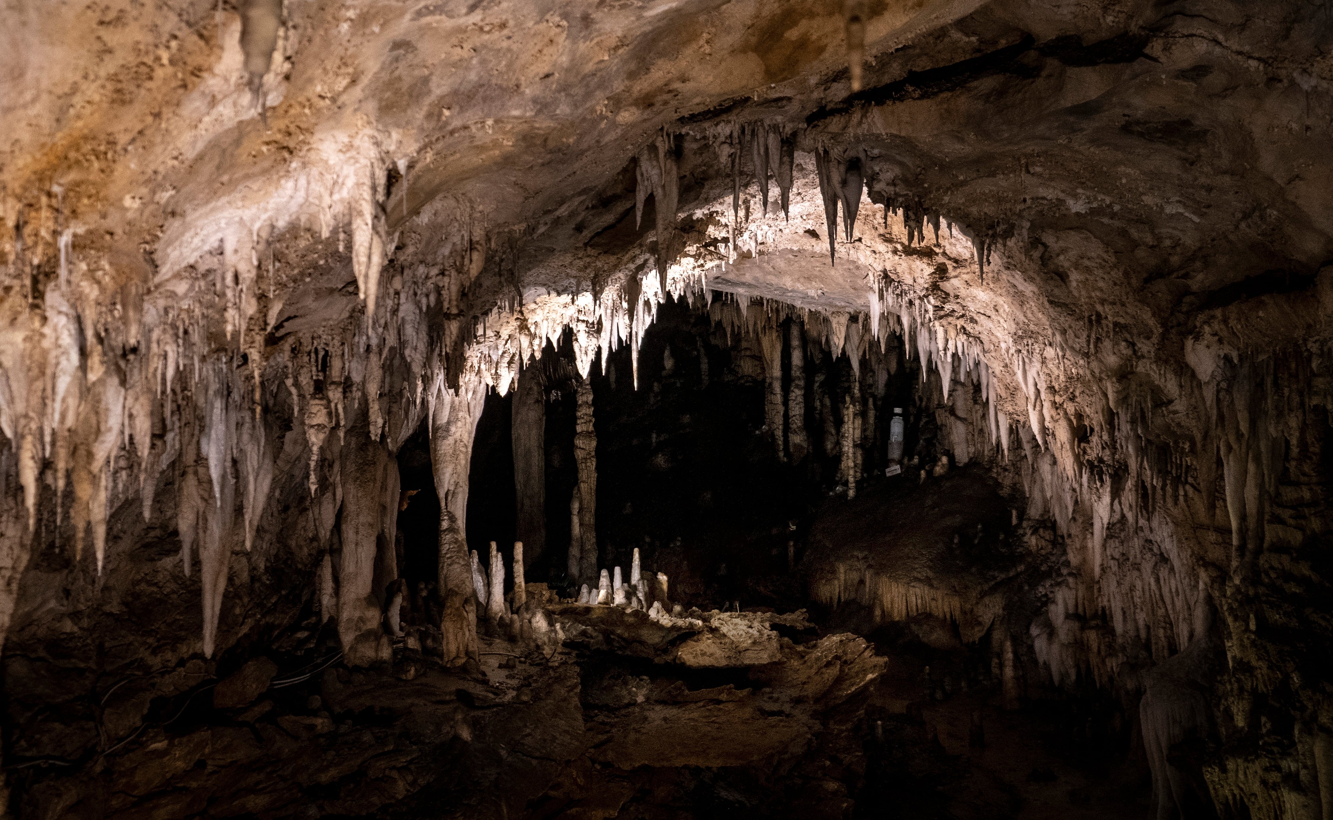 Interior de una cueva, caverna, con estalactitas y estalagmitas. La iluminación destaca las intrincadas formaciones y texturas de la roca, y crea una atmósfera misteriosa