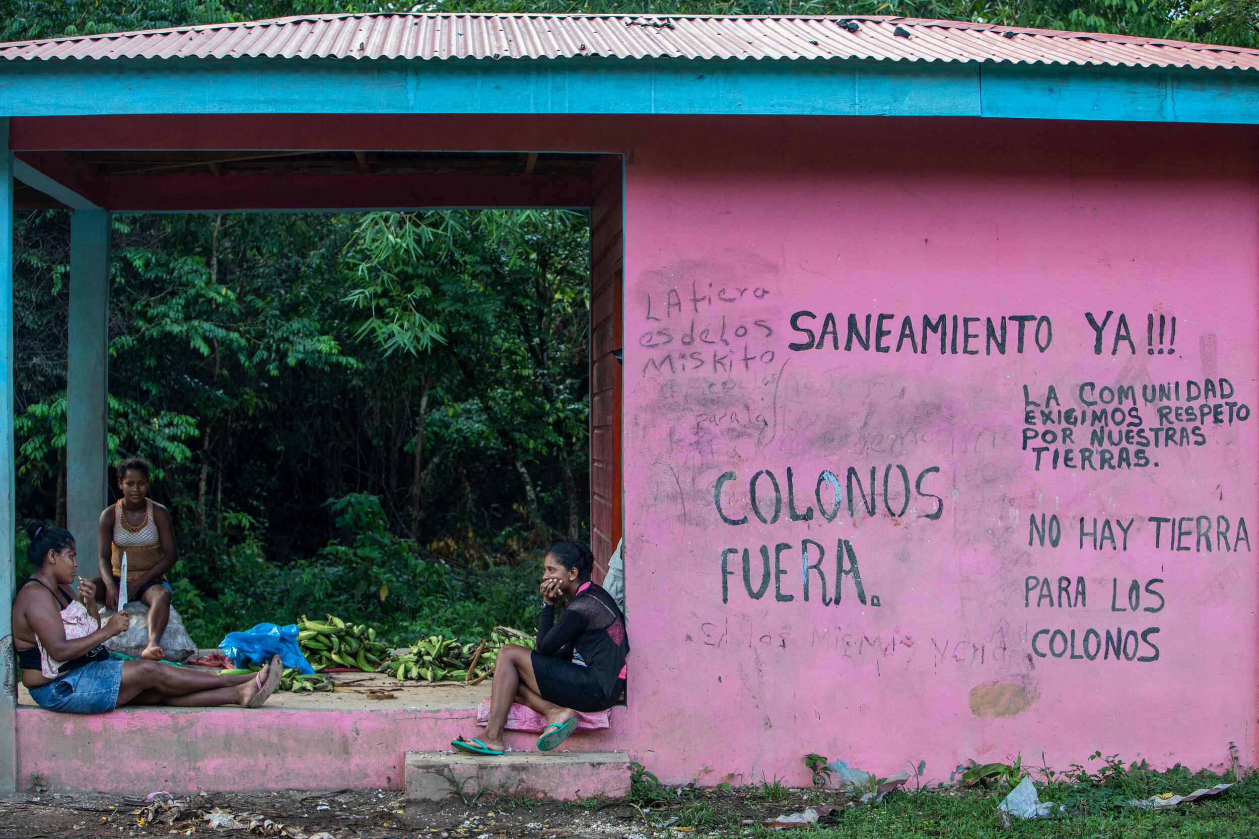 Mujeres indígenas miskitas sentadas junto a un muro con pintadas en las que se lee «Fuera colonos», «Saneamiento ya», «Nuestra comunidad exige respeto a nuestras tierras» y «No hay tierra para colonos», en la comunidad de Sangnilaya, Puerto Cabezas, Región Autónoma Costa Caribe Norte de Nicaragua, el 24 de septiembre de 2020.