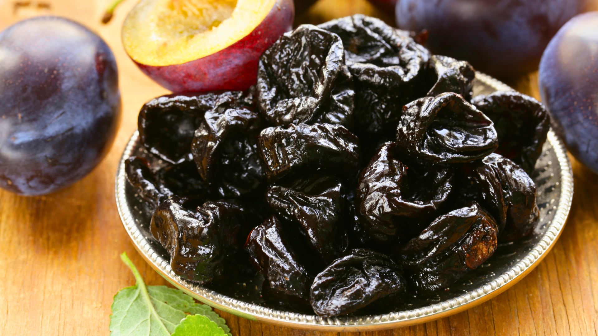 Plato metálico lleno de ciruelas pasas oscuras y brillantes, acompañado de ciruelas frescas enteras y cortadas; imagen que representa los beneficios nutricionales de este fruto seco para los huesos, el sistema cardiovascular y el control de la glucosa.