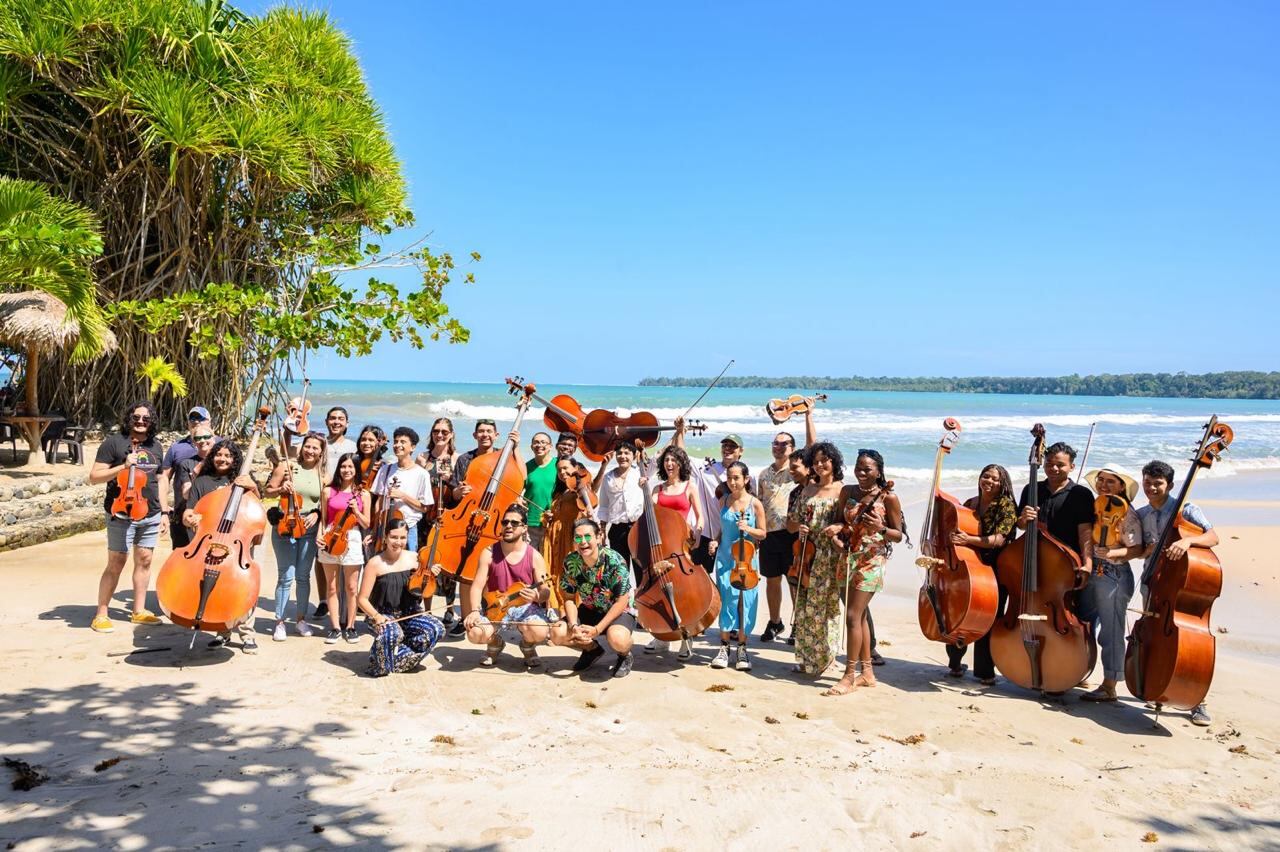 Imagen en la que se ven varios músicos con sus respectivos instrumentos como chelos, guitarras y violines posando frente a una playa.