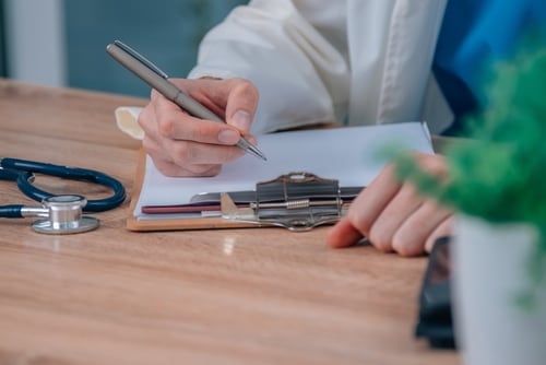 Mano de un médico escribiendo en un portapapeles con estetoscopio sobre el escritorio.
