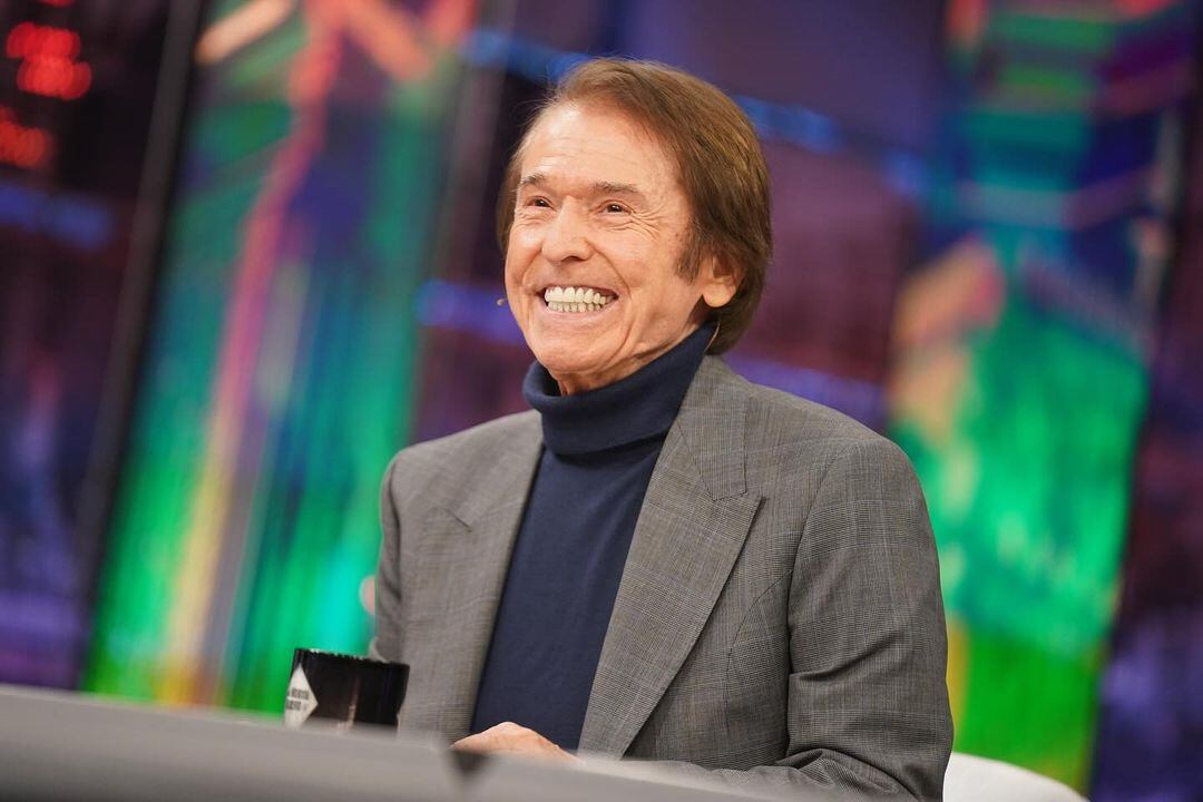 Imagen del cantante español Raphael sonriendo, usando un traje de color gris y camisa azul.