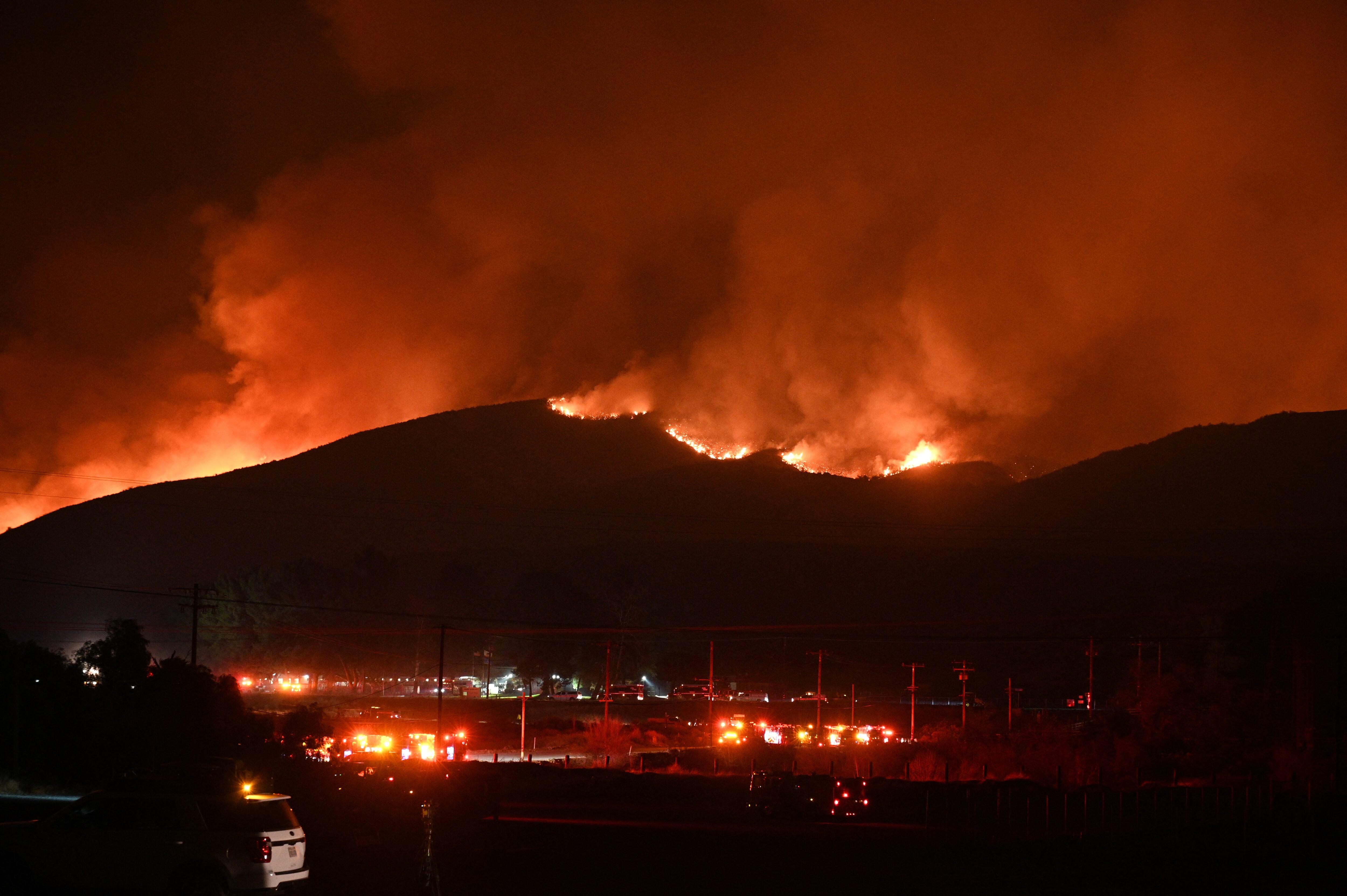 Camiones de bomberos y vehículos de seguridad pública se ven en primer plano mientras las llamas rojas y el humo se elevan desde el incendio Hughes en Castaic, en el noroeste del condado de Los Ángeles, California.