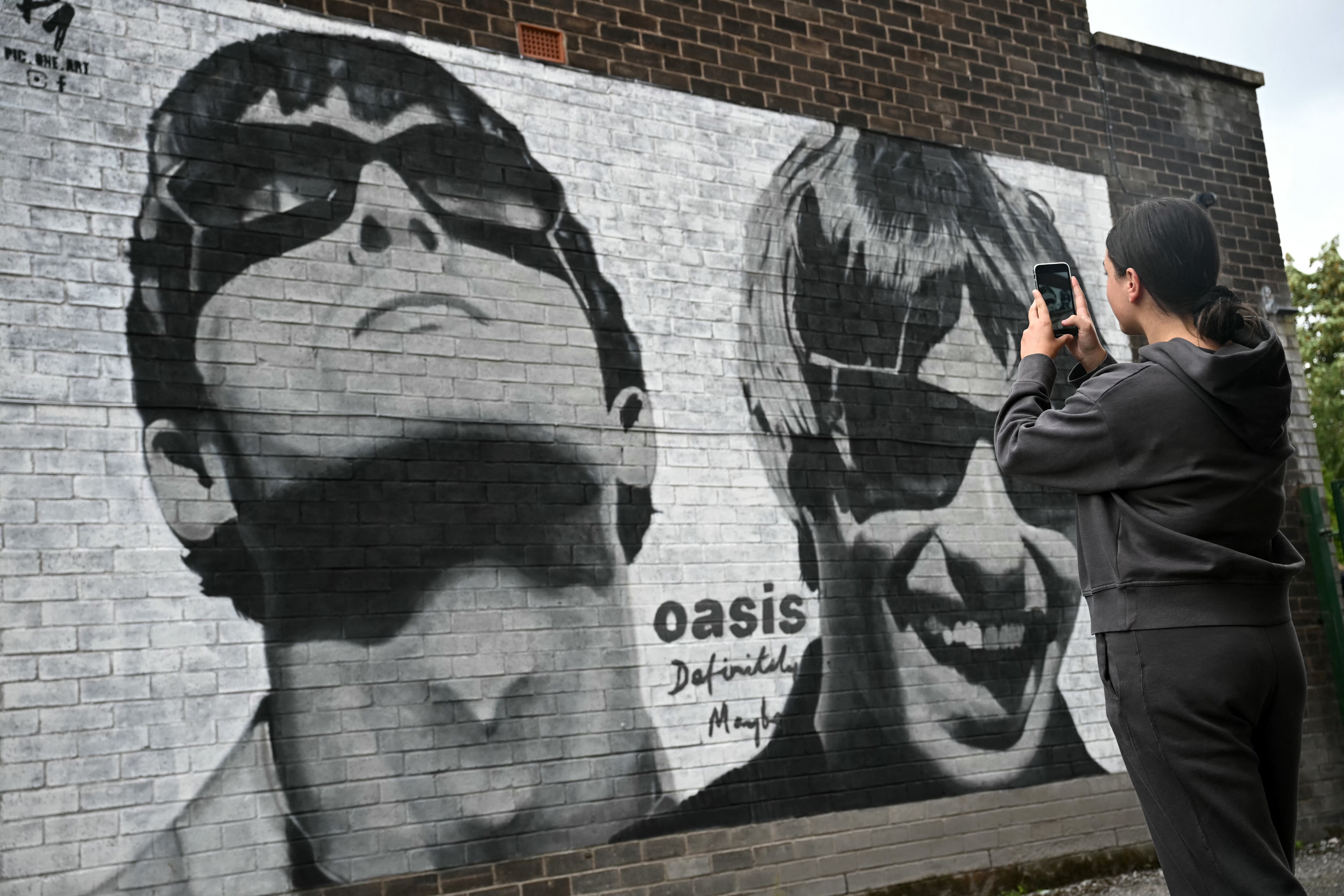 Fan Emily McShane, takes a photograph of a new street artwork depicting Oasis' Liam and Noel Gallagher, created by Manchester-based street artist Pic.One.Art. on the side of the Sifters Record store in Burnage, a suburb of Manchester, northern England on August 27, 2024. British rock legends Oasis announced Tuesday they will reunite for a worldwide tour, as brothers Liam and Noel Gallagher ended an infamous 15-year feud. The band behind hit songs including "Wonderwall", "Don't Look Back In Anger" and "Champagne Supernova", will play an initial 14 gigs next year in Cardiff, Manchester, London, Edinburgh and the Irish capital, Dublin, starting in July. (Photo by Paul ELLIS / AFP) / RESTRICTED TO EDITORIAL USE - MANDATORY MENTION OF THE ARTIST UPON PUBLICATION - TO ILLUSTRATE THE EVENT AS SPECIFIED IN THE CAPTION
