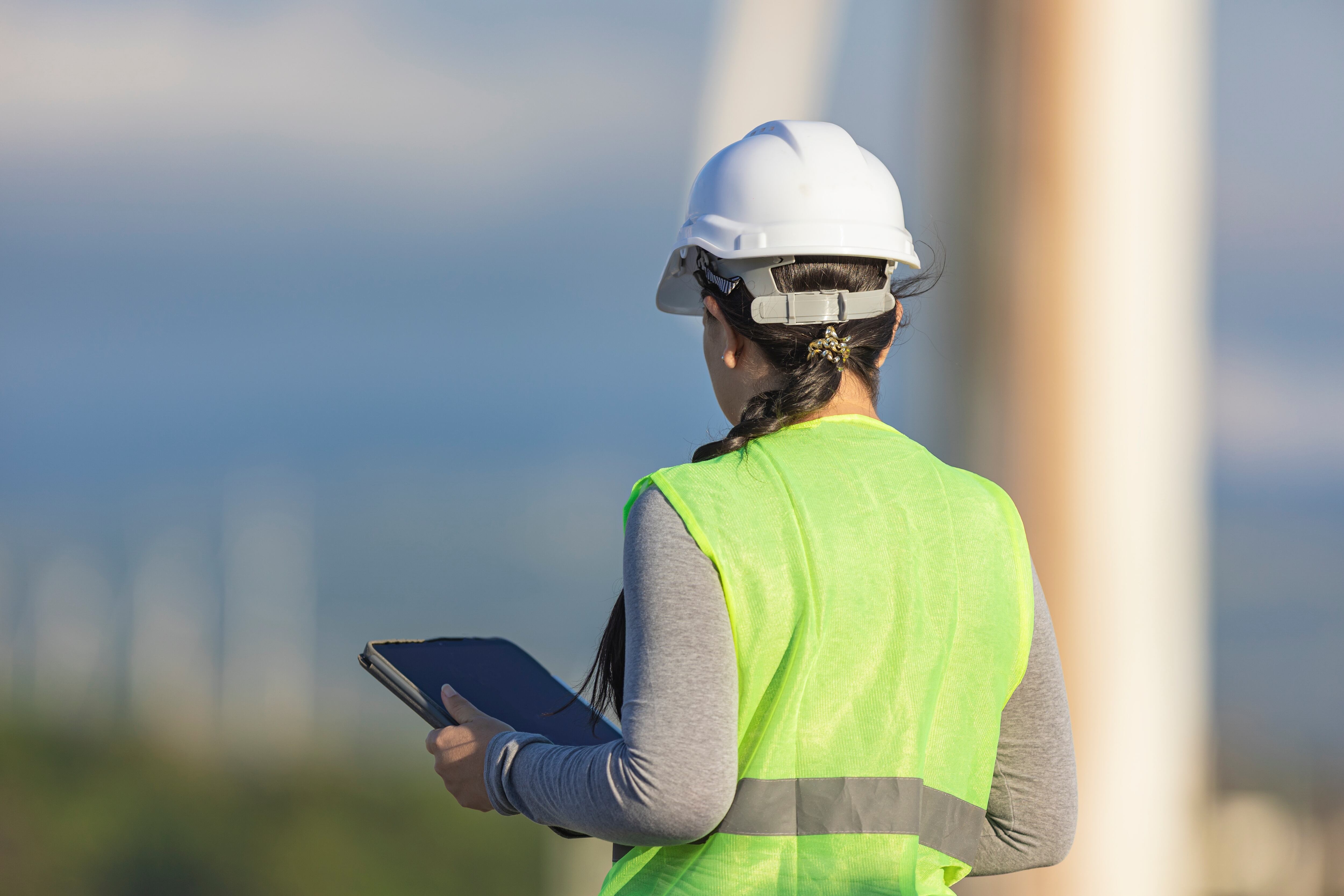 Ingeniera de espaldas, con casco y chaleco de seguridad, sosteniendo una tableta digital