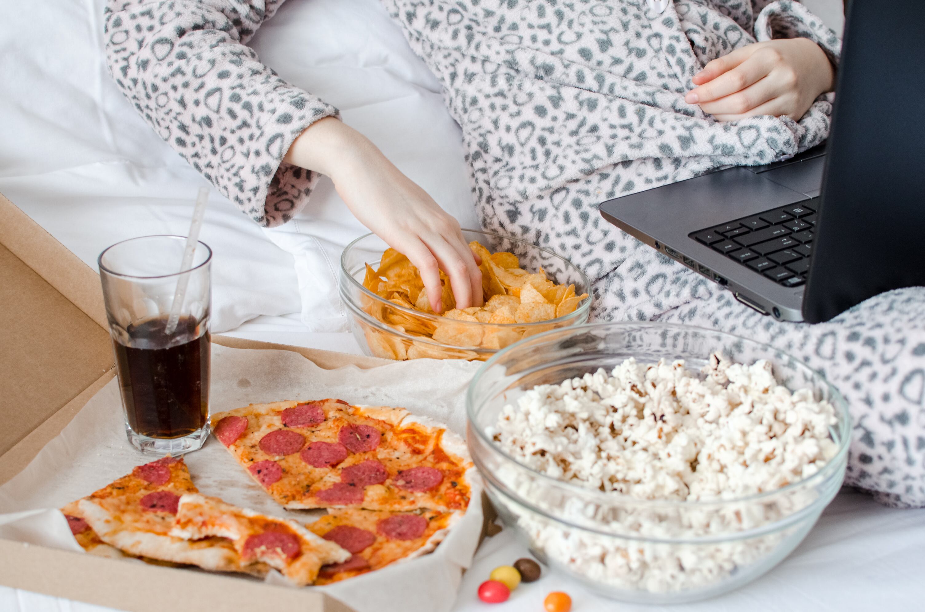 Una mujer joven a quien no se le ve la cara, acostada en una cama, con su laptop en los regazos y comiendo pizza, papas tostadas y popcorn con gaseosa.