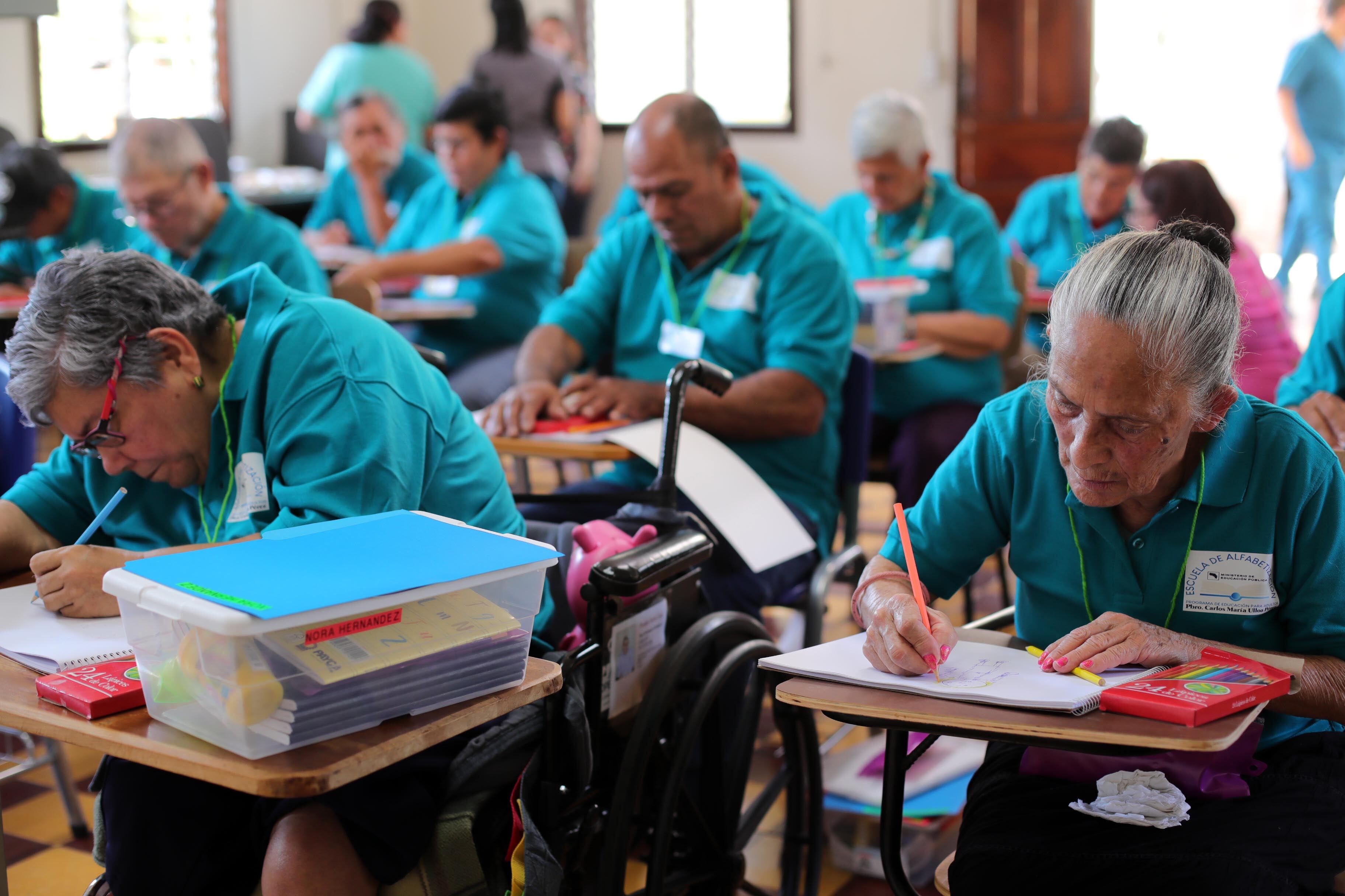 Clase en una escuela con varios adultos mayores sentados en pupitre, con camiseta turquesa. En su pupitre tienen los útiles escolares.