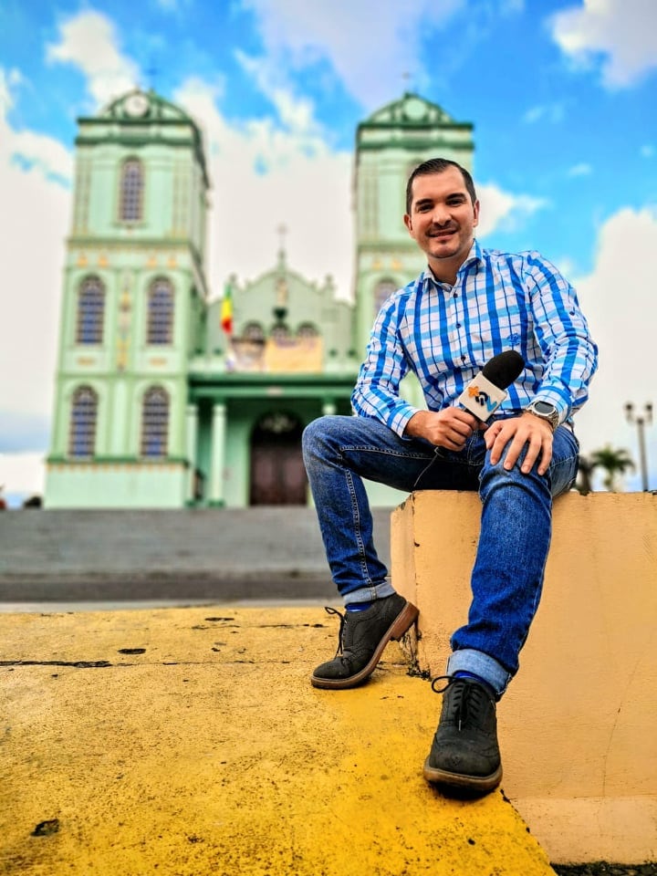 Imagen del periodista José Ernesto Herrera de 'Más que noticias' sentado frente a una iglesia y sosteniendo el micrófono del programa en su mano derecha. Usa camisa a cuadros azul y un pantalón de mezclilla.