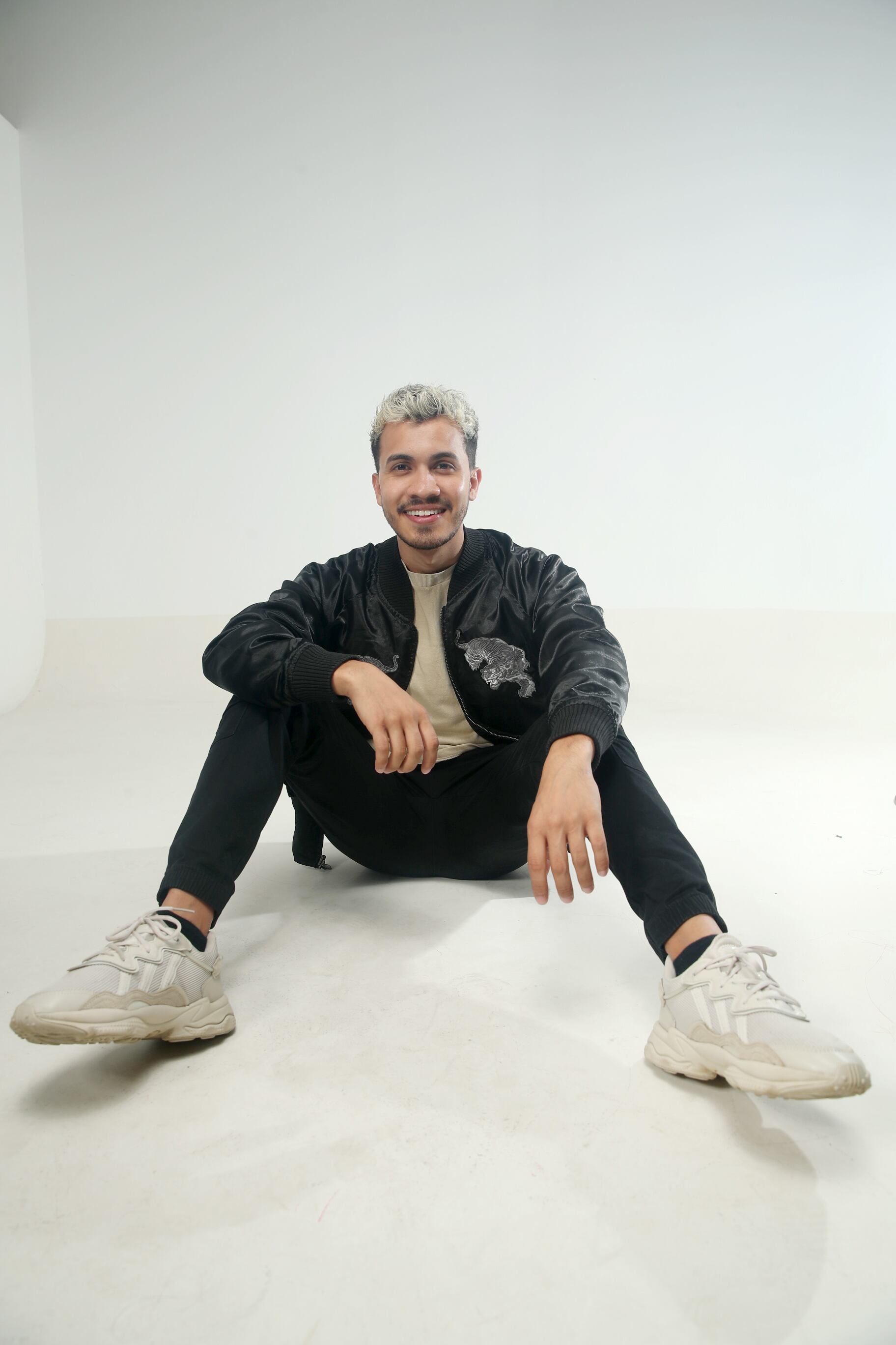 El youtuber Araya Vlogs posando sentado en el suelo, usa chaqueta y pantalón negro y zapatos blancos.
