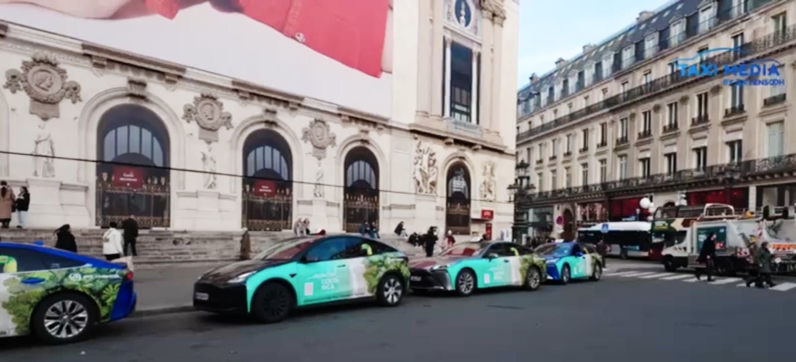 Parada de taxis en Paris. Se ven a la orilla de la acera, entre edificios antiguos de color beige, que no sobrepasan los tres pisos. Los taxis son bnacos con imágenes de bosques tropicales en su carrocería.