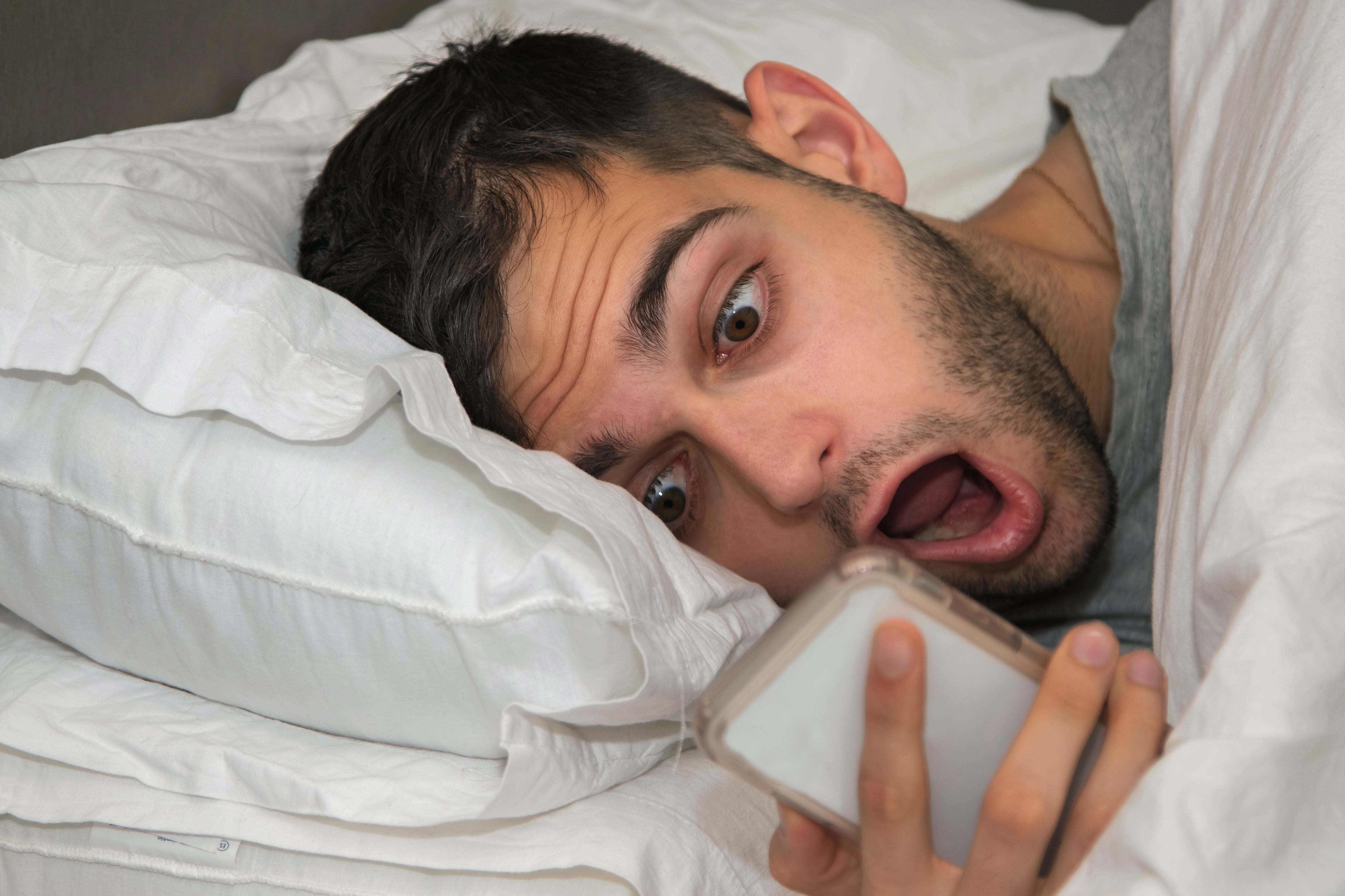Hombre viendo su celular en la cama, sobre la almohada, con expresión de terror, asustado, miedo, impresión, sorpresa