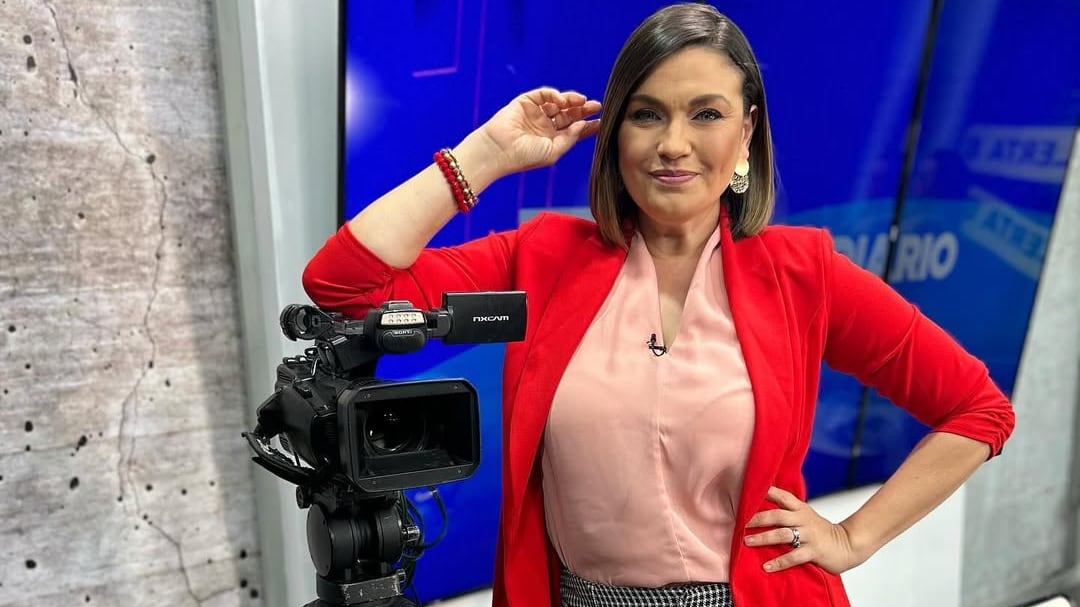 Foto en primer plano de la periodista Natalia García, usa un saco rojo, una blusa rosada y tiene su brazo sobre una cámara de televisión.