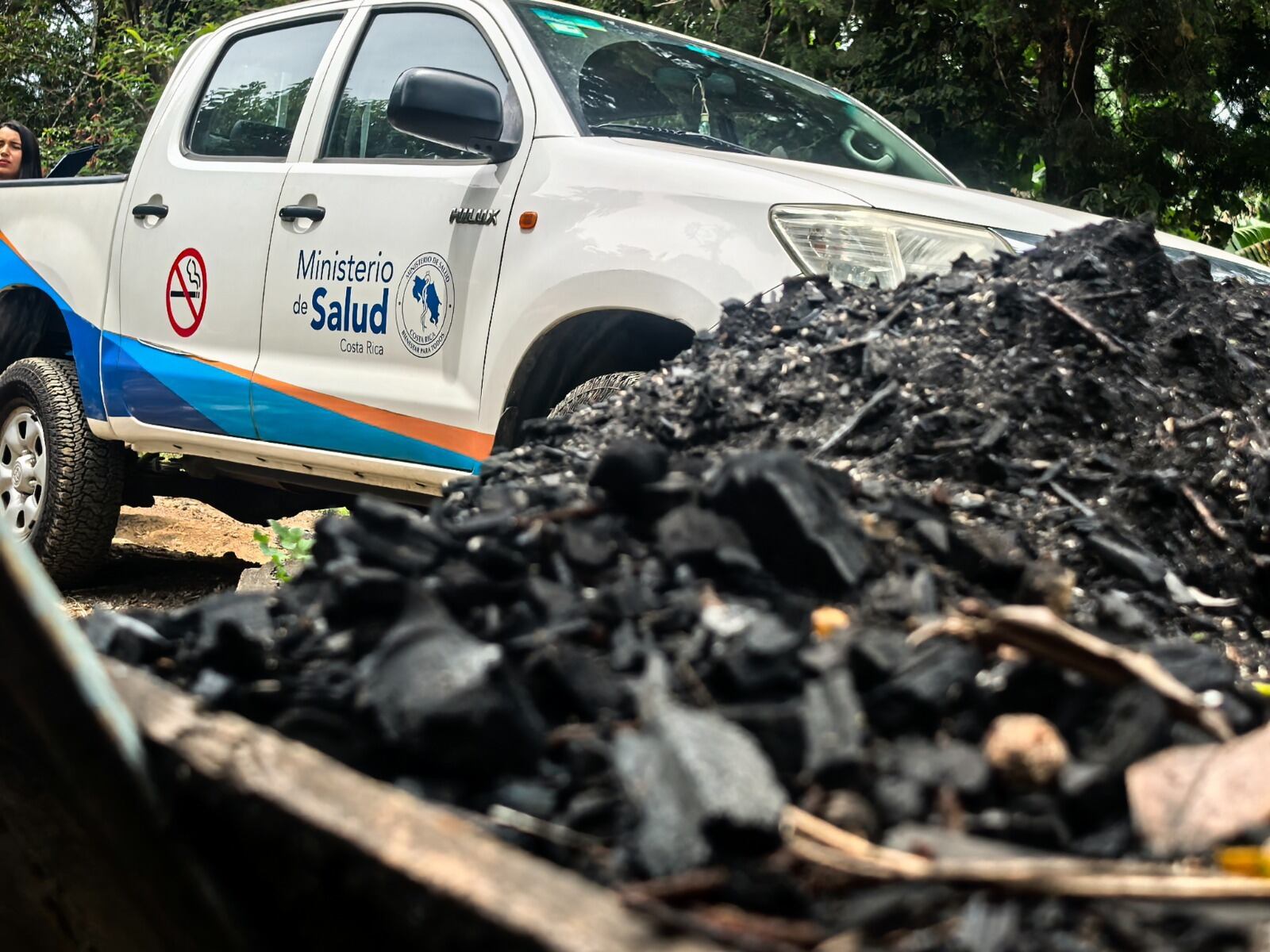Una montaña de carbón en primer plano. Al fondo, un vehículo del Ministerio de Salud.