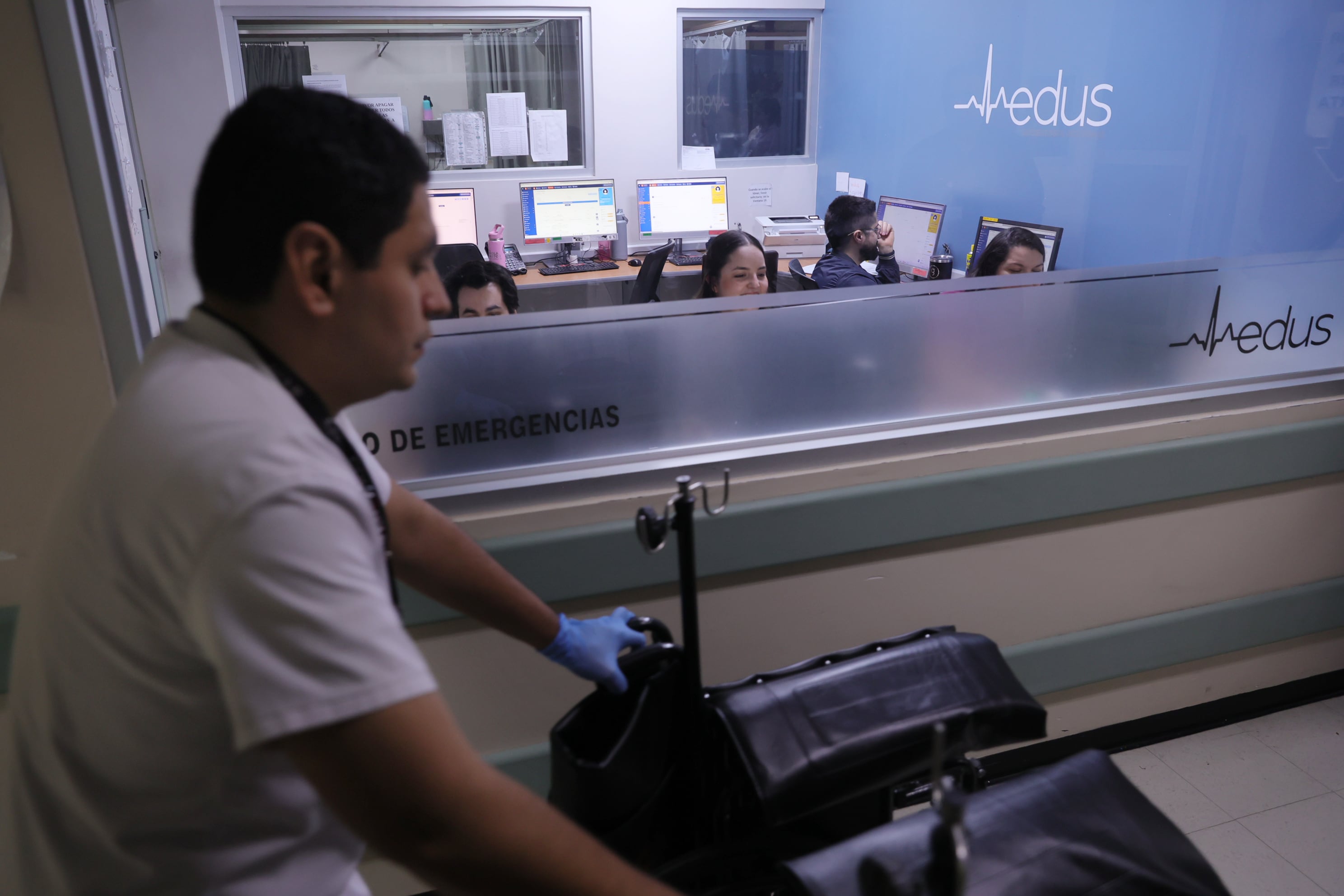 Hombre vestido con camisa blanca empuja una silla de ruedas vacía. Al fondo, tras un ventanal, se ve un grupo de gente frente a varias computadoras.