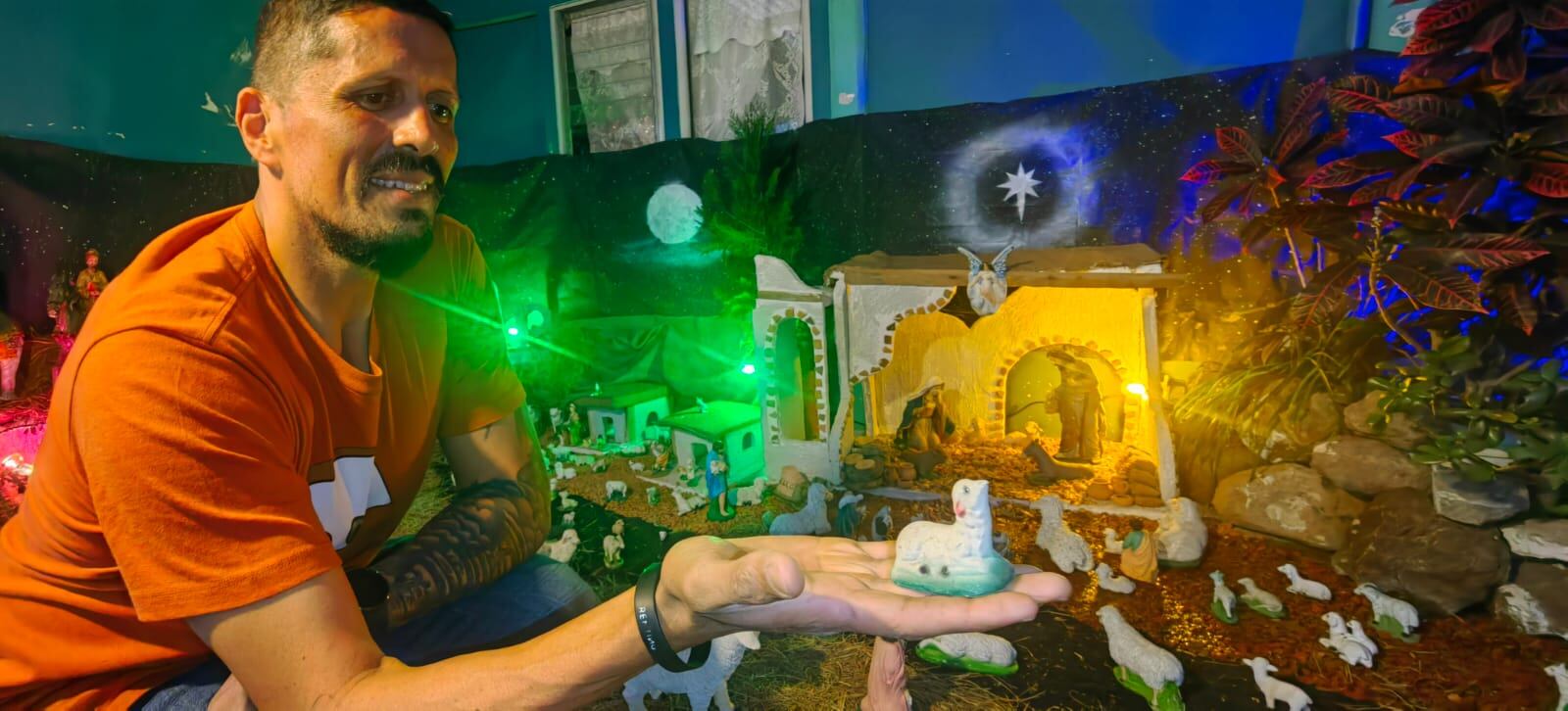 Hombre acuclillado en un portal, en su mano extendida tiene la figura de una oveja. Al fondo, se observa el pesebre.