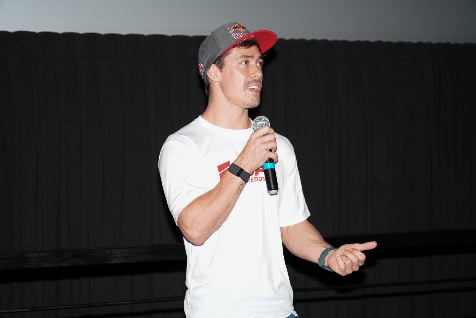 El ciclista Kenneth Tencio en una conferencia de prensa. Sostiene un micrófono, usa gorra gris y camiseta blanca.