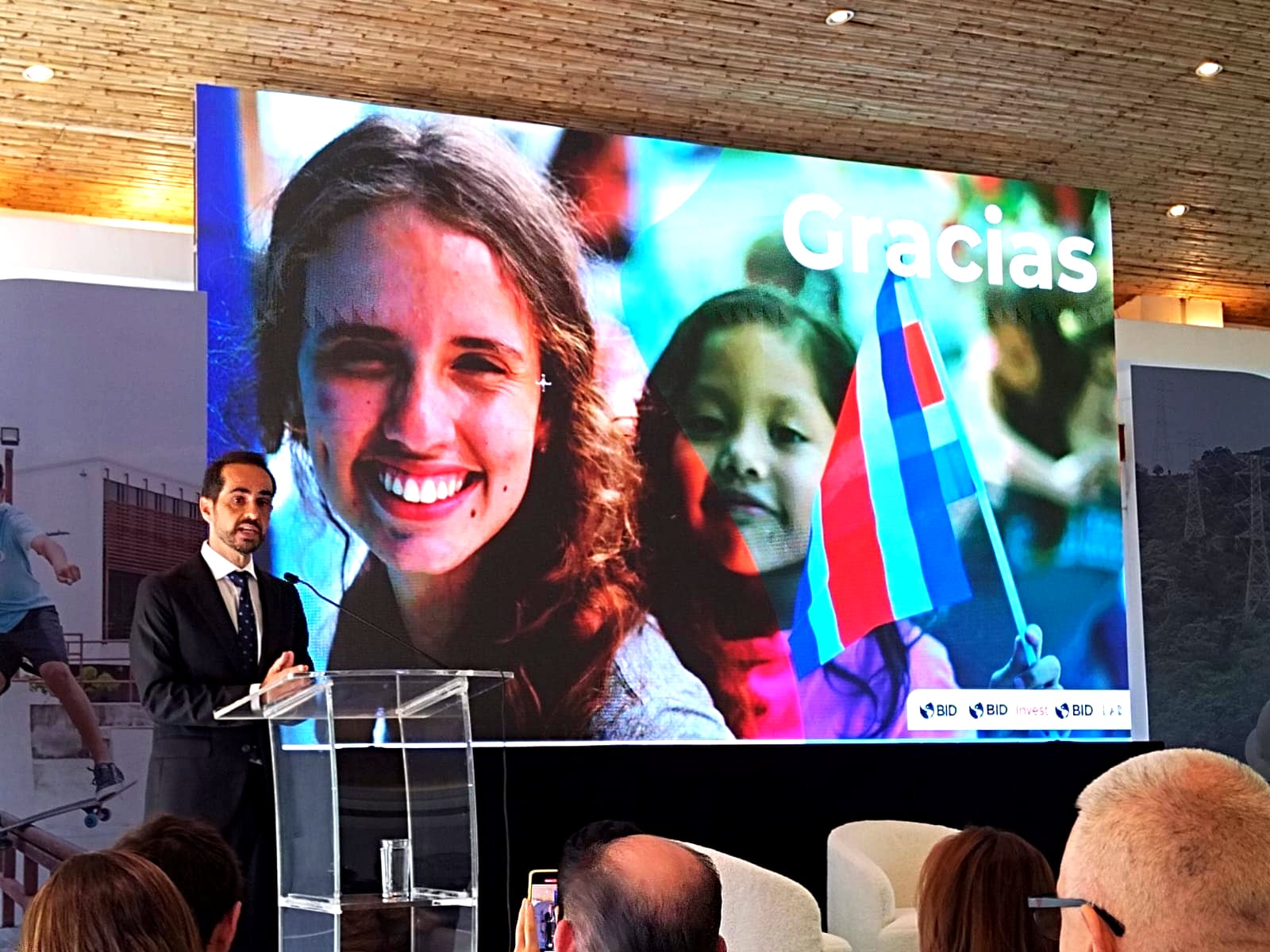 Hombre de traje hablando en un auditorio, tras él una pantalla con un una mujer sonriendo, una niña y la bandera de Costa Rica