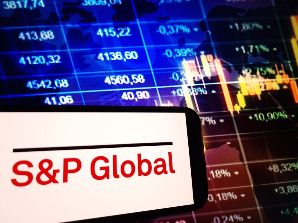 Logo de S&P Global en pantalla de celular, con gráficos de mercado financiero en segundo plano, relacionado con la calificación crediticia y la evaluación económica de Colombia.