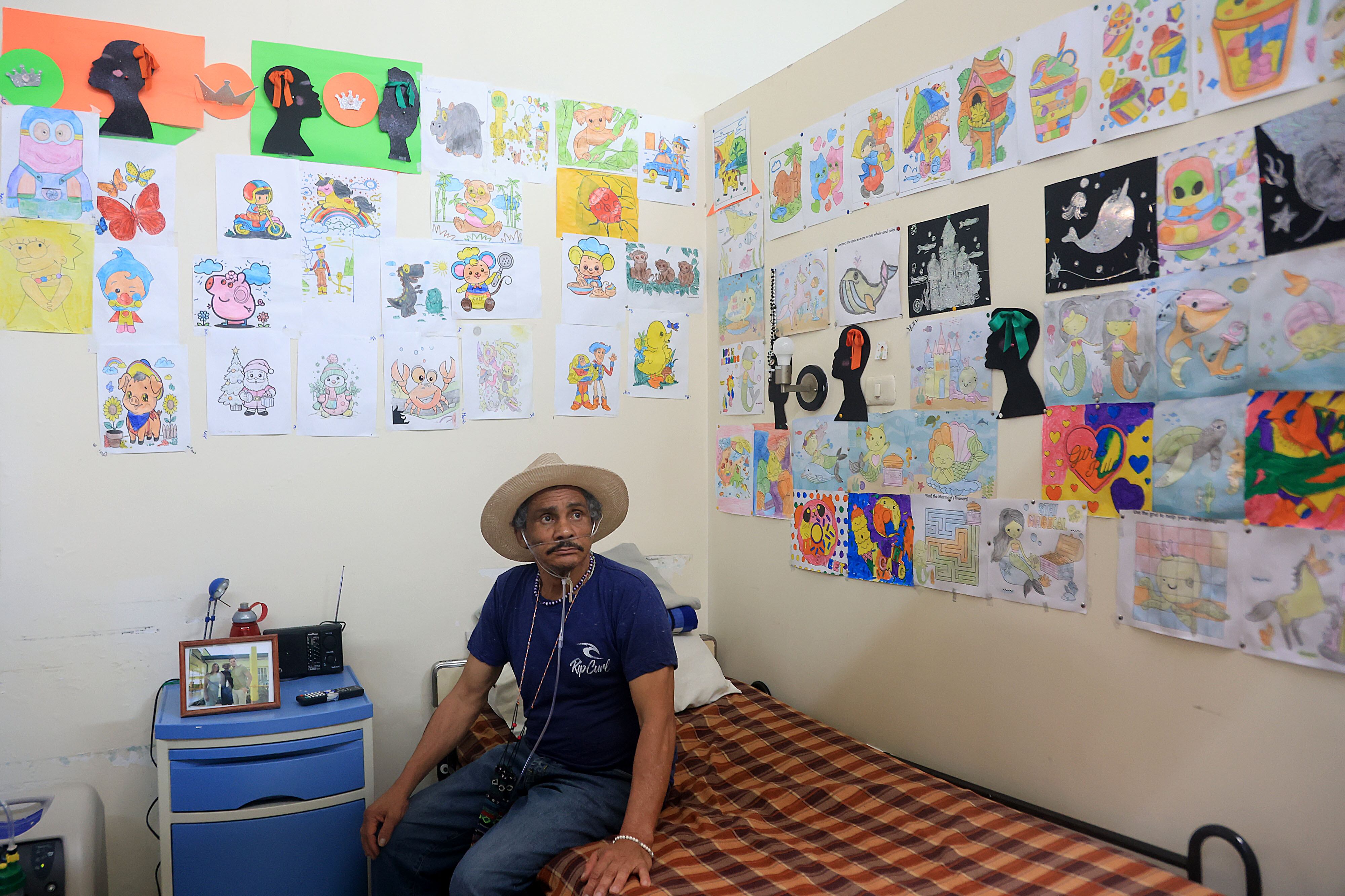 Hombre sentado en camastro en un cuarto con paredes blancas. El hombre viste camiseta azul y jeans, Lleva un sombrero de ala ancha. Tiene bigote. En las paredes hay muchos dibujos pintados en papel.