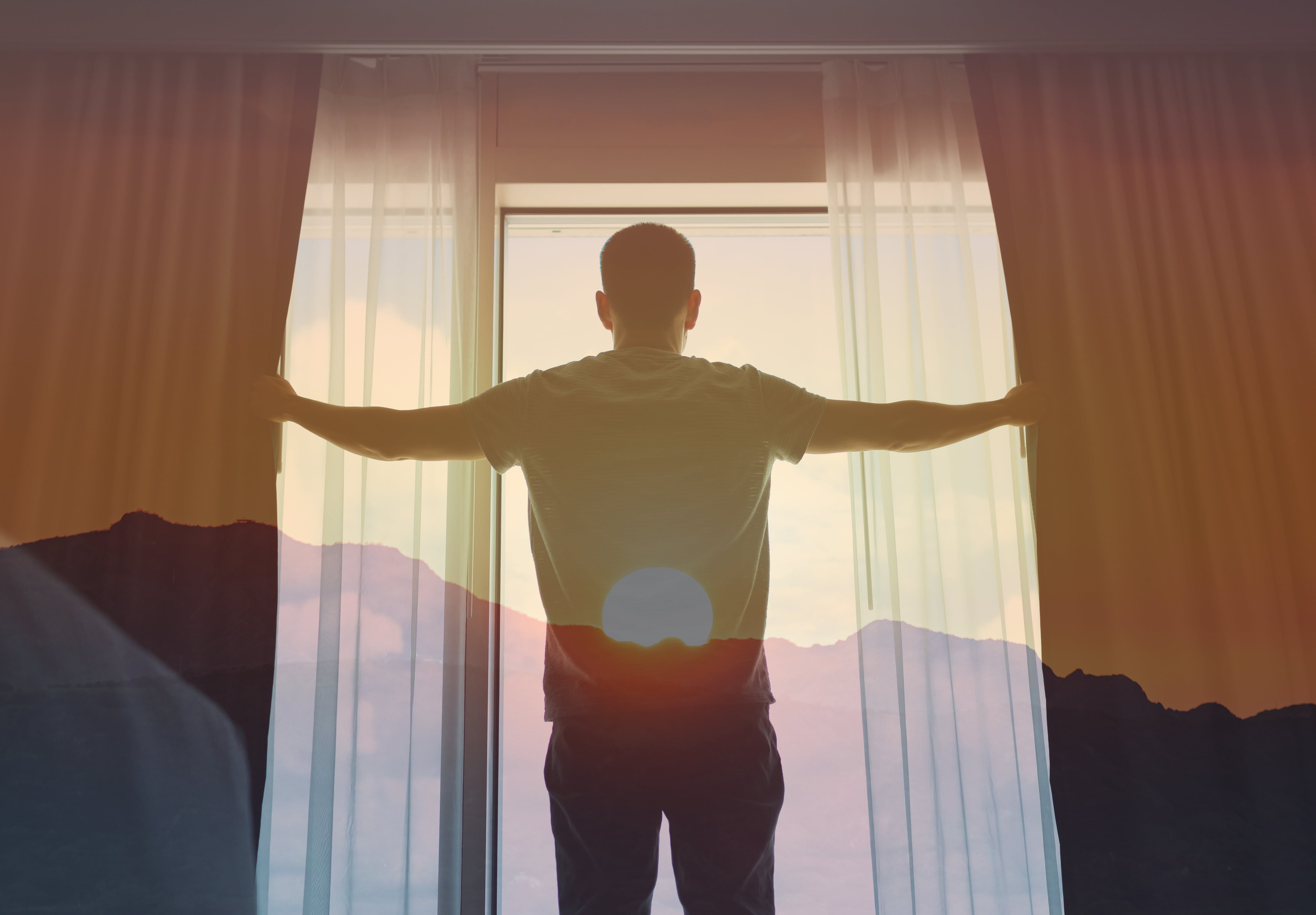 Hombre de espaldas, mirando el amanecer a través de la ventana de su habitación, montañas al fondo y el sol saliendo