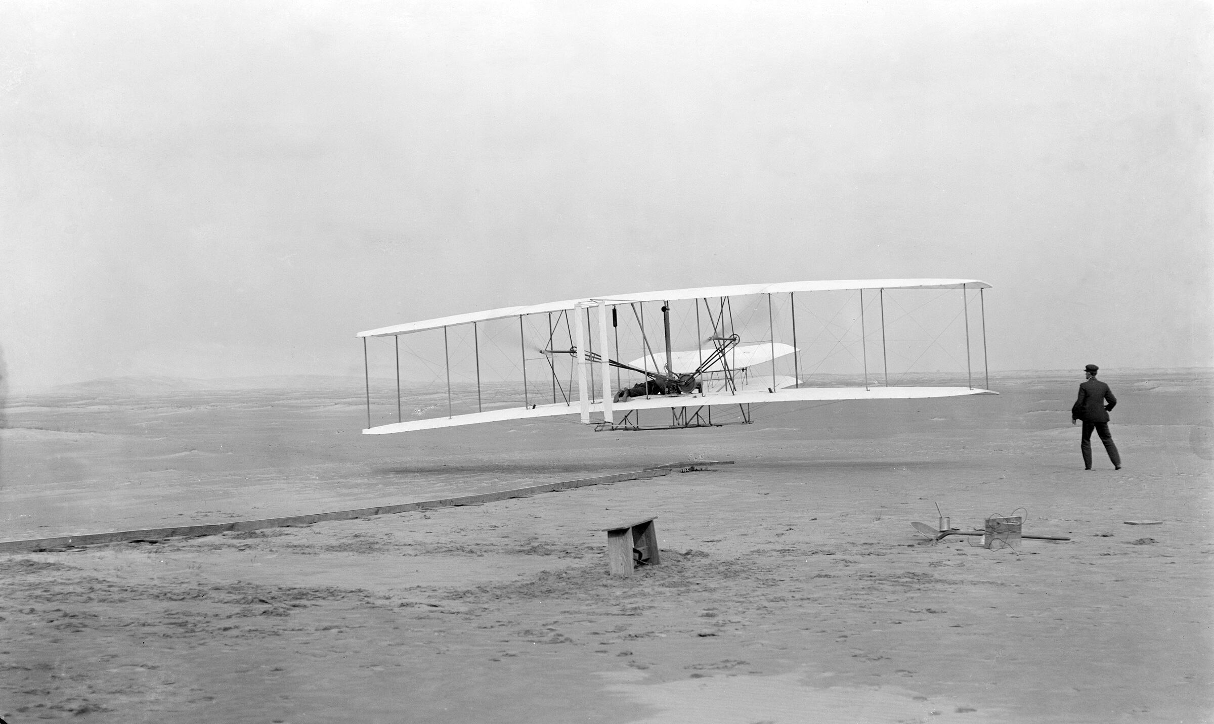 El aeroplano Wright Flyer durante su primer vuelo controlado en 1903, con Orville Wright a bordo y Wilbur Wright observando en tierra, en Kitty Hawk, Carolina del Norte.
