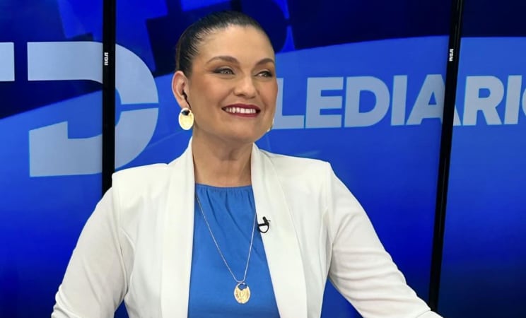 Foto en primer plano de la periodista Natalia García con el fondo de Telediario, usa un saco blanco, blusa azul y está sonriendo.